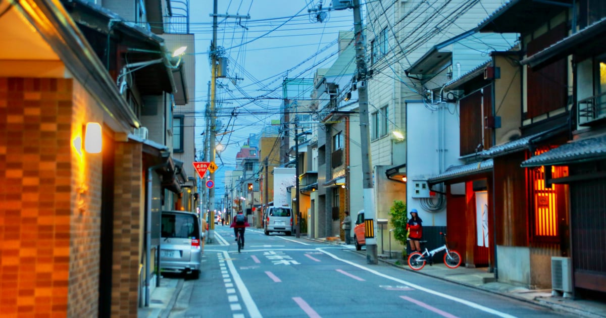 Quiet neighbourhood street in Kyoto at dusk, with wooden townhouses, small shops and a cyclist riding past